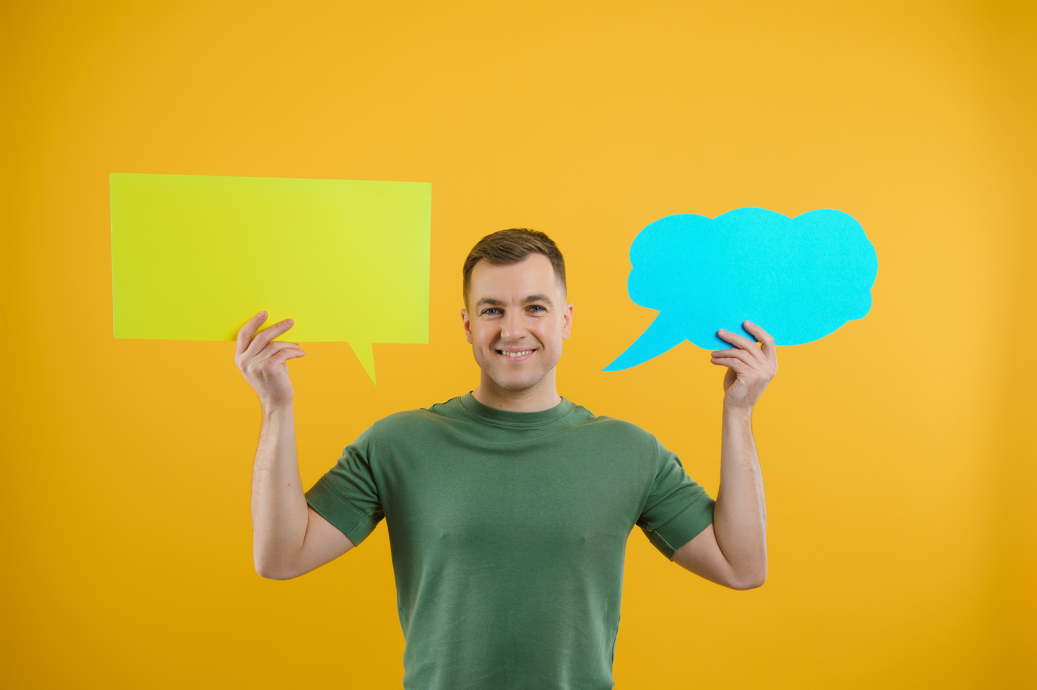 Portrait of cheerful smiling young handsome man holding and looking up at speech bubble with empty space for text on colorful yellow studio background